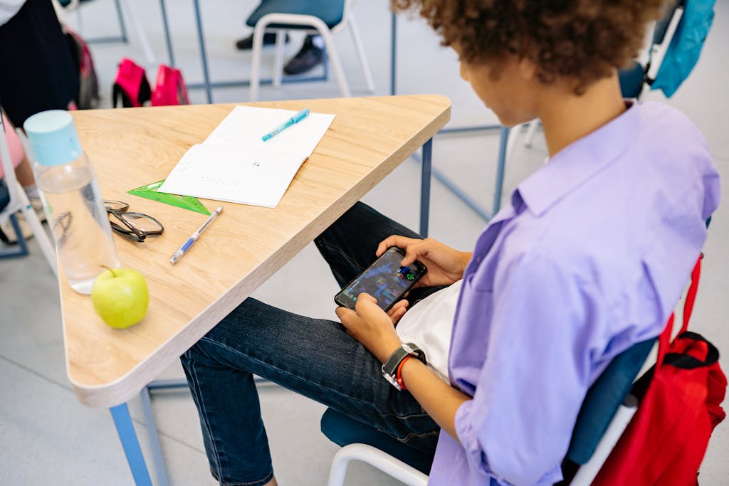 A child playing a mobile game at a school desk with school supplies nearby, captured indoors during class.