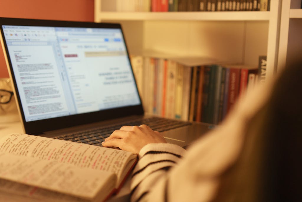 A person studying online with a laptop and open book in a cozy home setting, surrounded by shelves of books.