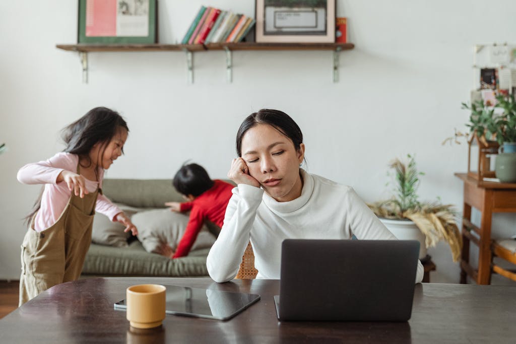A tired mother working remotely on her laptop while children play in the background.