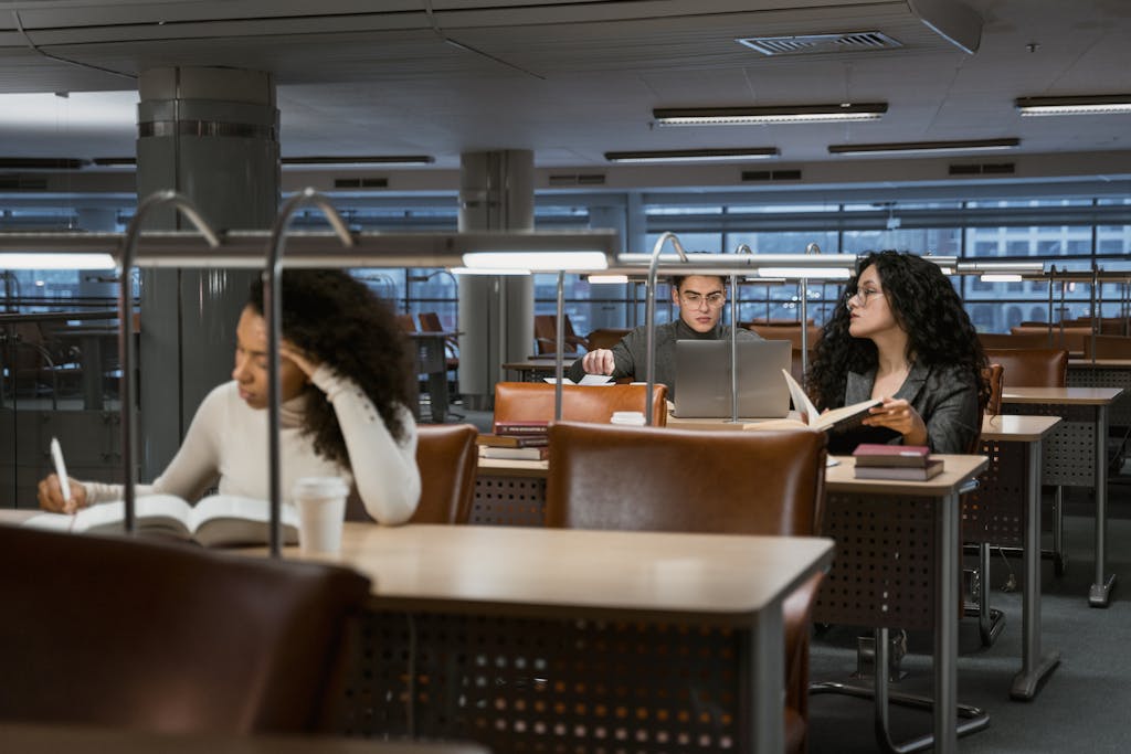 College students studying in a modern library setting, concentrating on books and laptops.