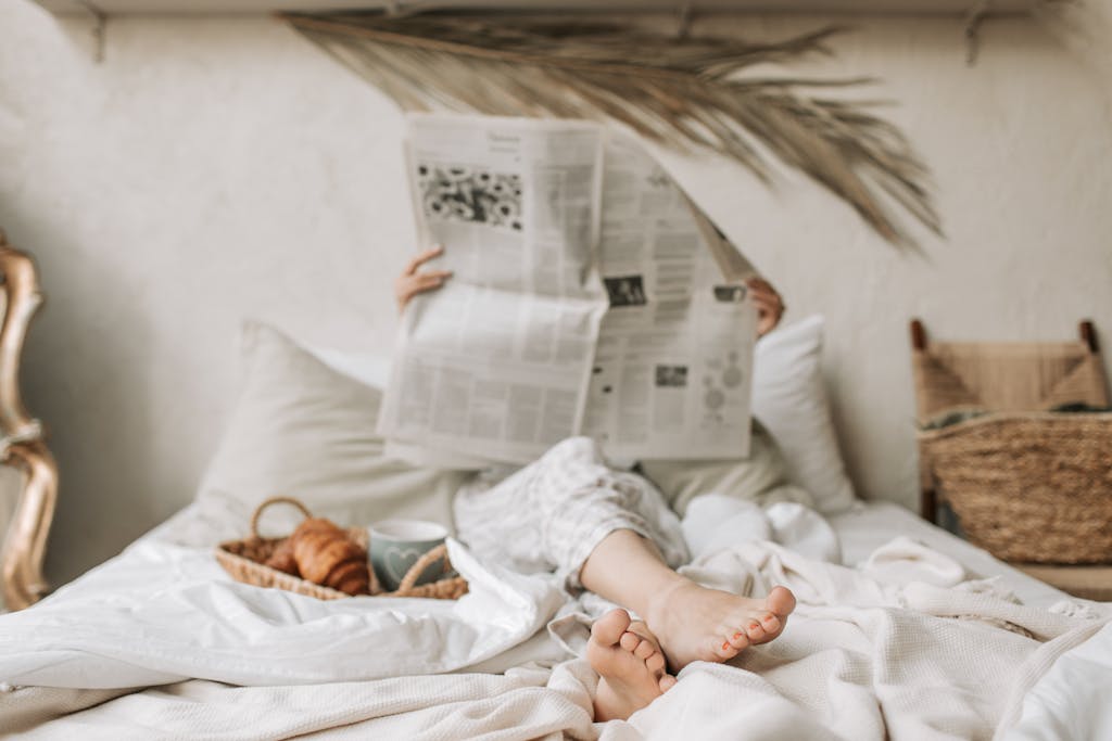 Person relaxing in bed with a newspaper, coffee, and croissant for a calm morning vibe.