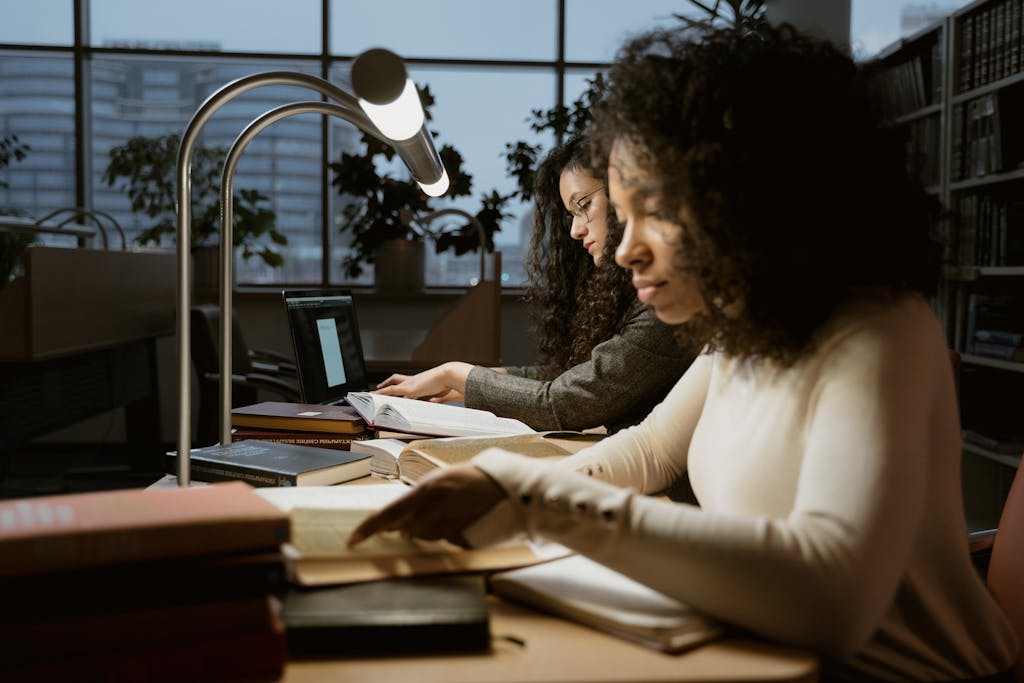 Two women intensely studying in a library setting, under warm desk lamps.