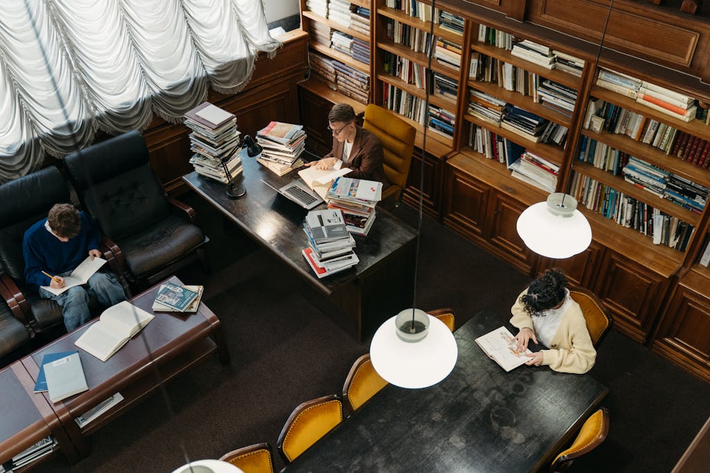 University library with students studying at desks, surrounded by bookshelves and ceiling lights.