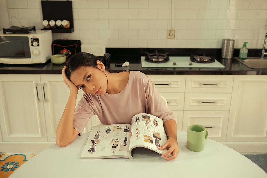 Woman sitting in a kitchen, reading a magazine at a table with a thoughtful expression.