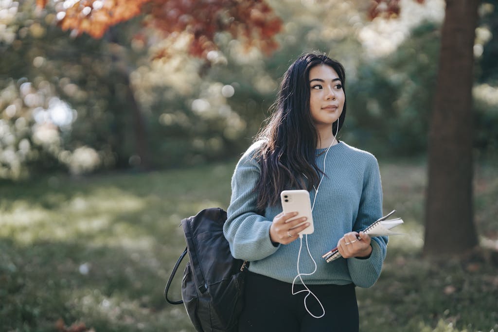 A young woman enjoys music through earphones while holding a smartphone and notebook in a lush autumn park.