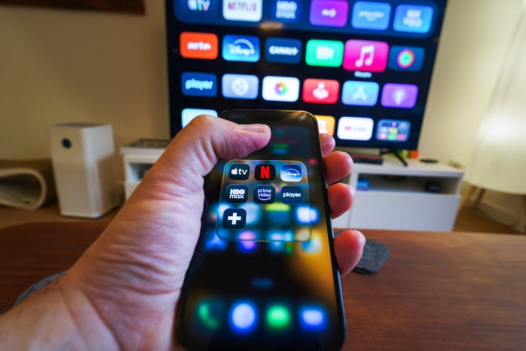 Close-up of a hand holding a phone displaying streaming apps in front of a TV with multiple app icons.