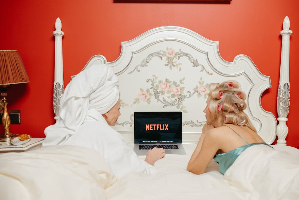 Two women lying in bed watching Netflix on a laptop, creating a cozy and bonding atmosphere.