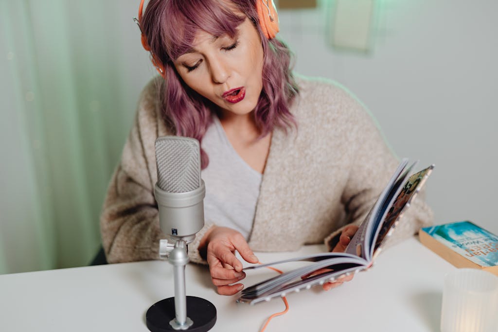 Woman with pink hair singing while reading a book, wearing headphones, seated at a white table with a microphone.