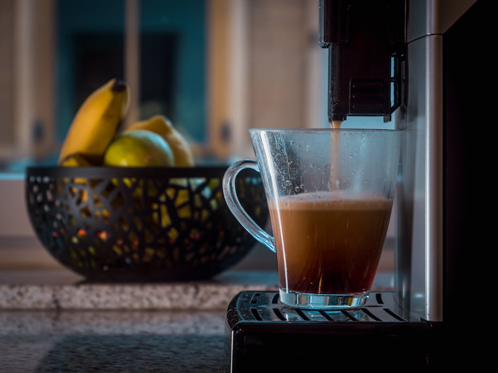 Glass cup catching freshly brewed coffee near a bowl of bananas and apples in a modern kitchen.