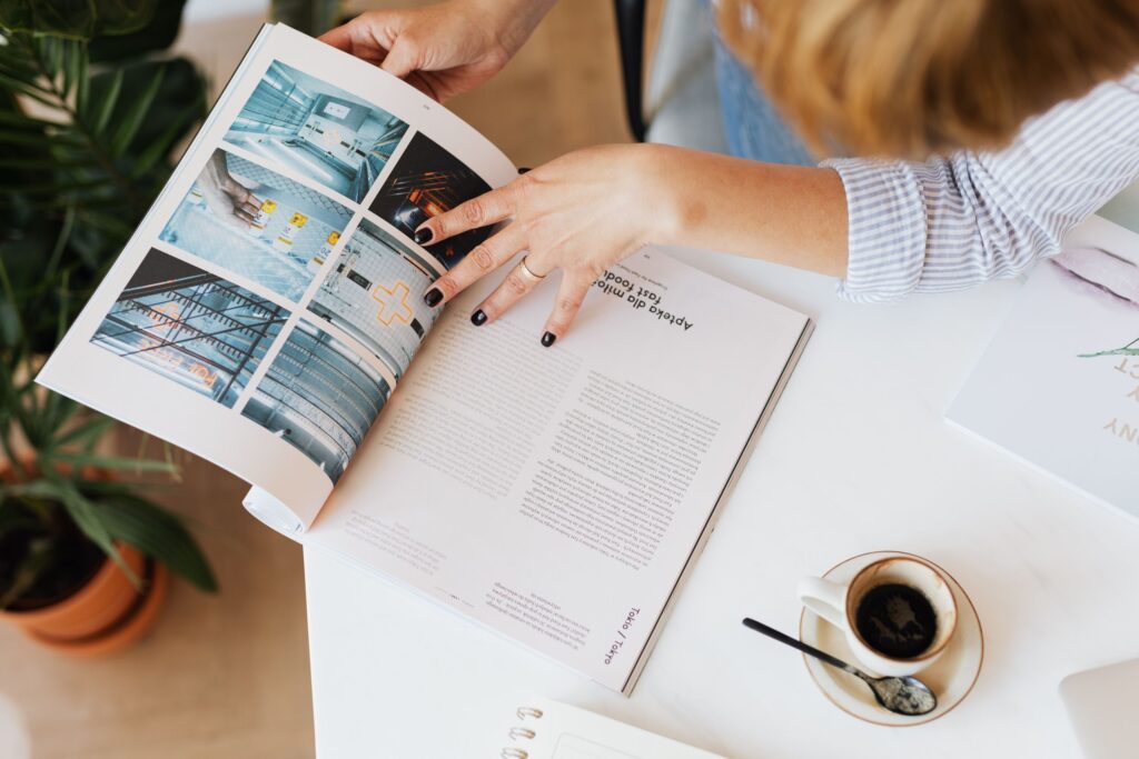 From above crop anonymous female sitting at desk with cup of aromatic americano and reading interesting magazine article near green plant at home office
