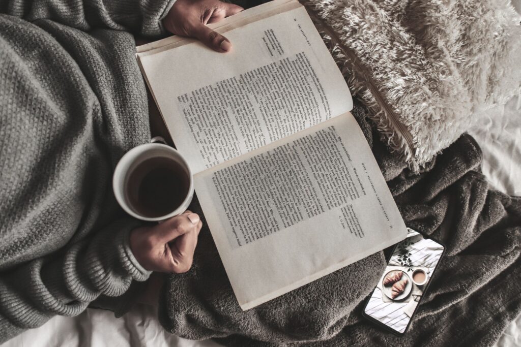 From above of crop anonymous female with mug of hot tea reading book under plaid near smartphone at home