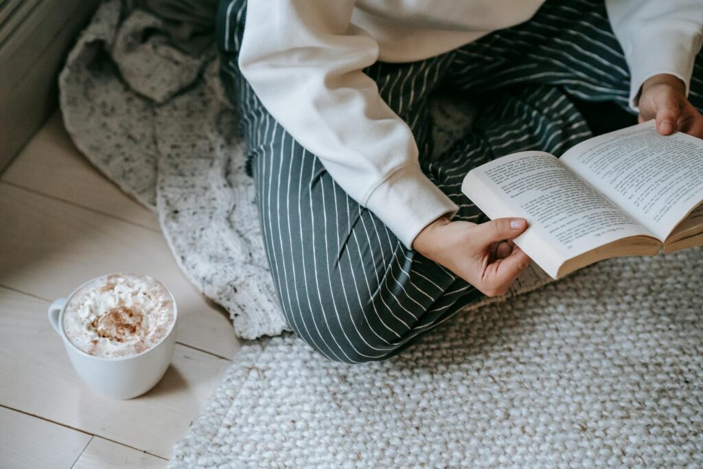 High angle of crop anonymous woman sitting on soft blanket on floor and reading literature with mug of coffee