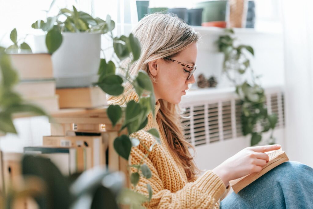 Side view of focused young lady with long blond hair in knitted sweater and eyeglasses reading novel sitting on floor near bookshelf in light apartment