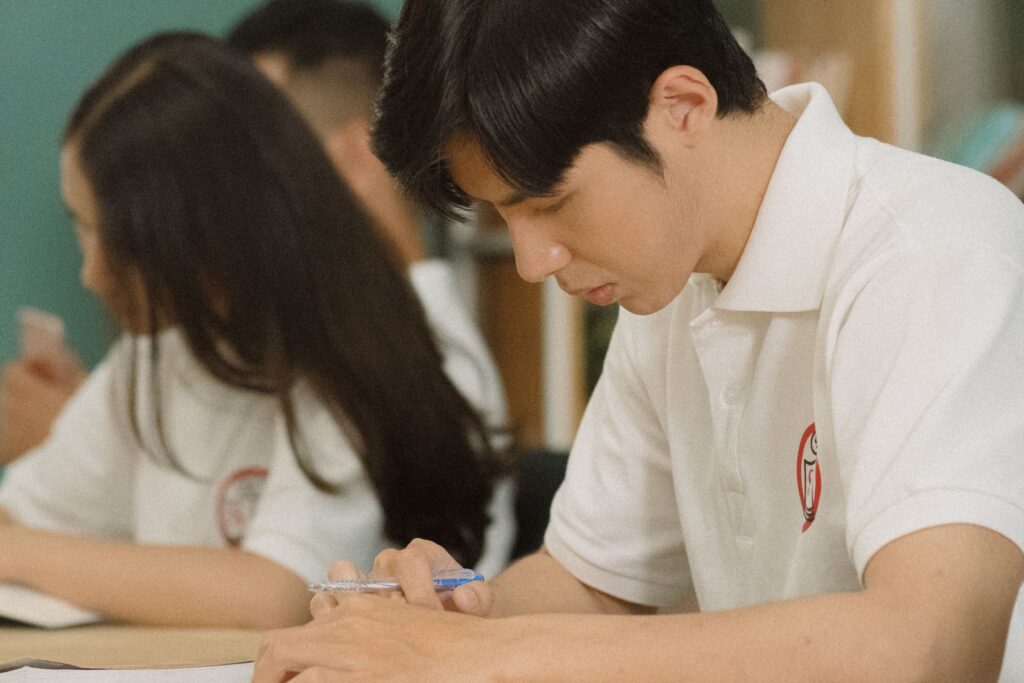 Two students focused writing in a classroom, wearing white polo shirts, adding notes with pens.