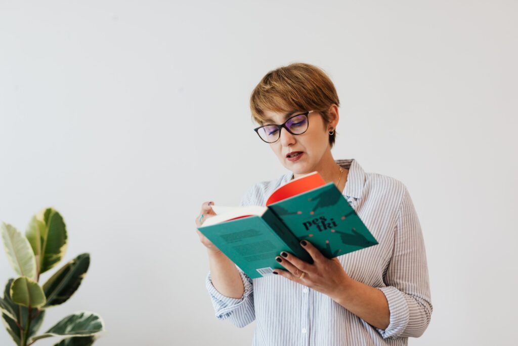Concentrated female in casual shirt and eyeglasses reading interesting book with green hardcover while standing against white wall and houseplant